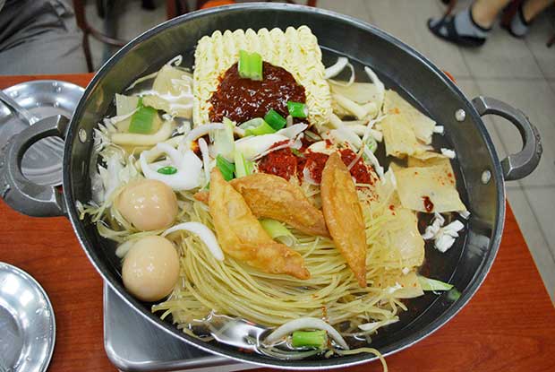 A metal pan, containing two types of noodles, a broth, some vegetables and meat, ready for cooking on a table top stove.