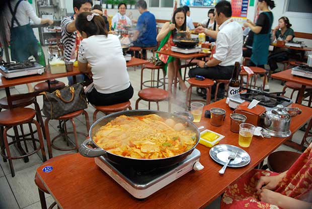 A crowded restaurant, in the background, people sitting on wooden stools eating, in the foreground a metal pan on a table top stove, simmering with noodles, meat, vegetables and sauce, the traditional ddeokbokki.