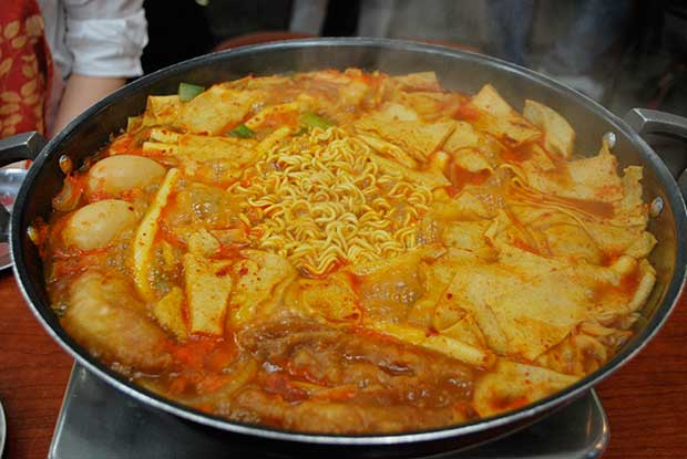 A close up of a metal pan with simmering ddeokbokki, a traditional Korean dish of noodles, vegetables, sauces and meat.