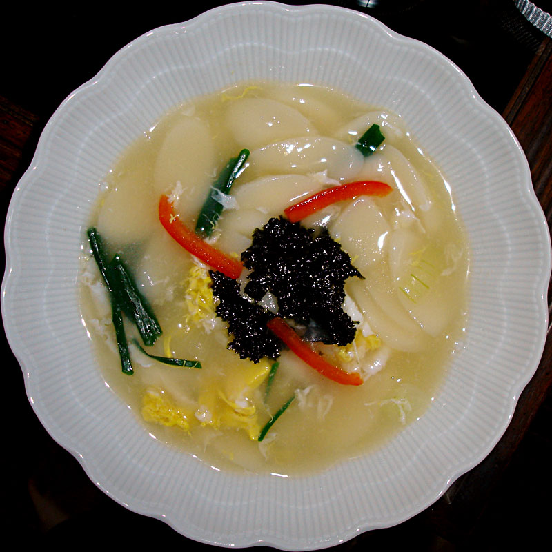 A Korean dish, Ddeokguk, a popular rice cake soup pictured in a white bowl with red and green chilies in a broth.