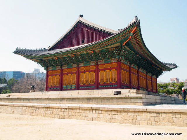 Exterior view of Jeonghwangeon throne hall. An ornate building in red, orange and green stands on a stone platform. In place of windows is orange fretwork, and ornate carving in the eaves.