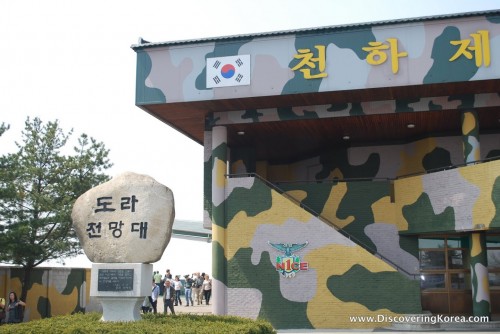 Outside of the Dora Observatory, painted in camouflage, with a sign on the left painted on a rock. Pedestrians milling in the background.