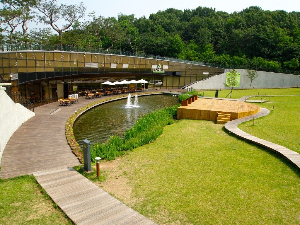 A semicircular brown building, with a wooden deck in front and a water feature with fountain. Umbrellas and cafe chairs outside, with the forest behind and lawns to the right of the frame.