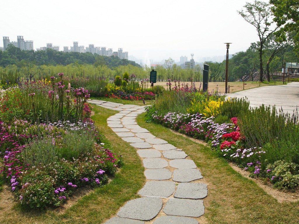 A stone pathway winds through beds of colorful flowers, with views of the city in the background.