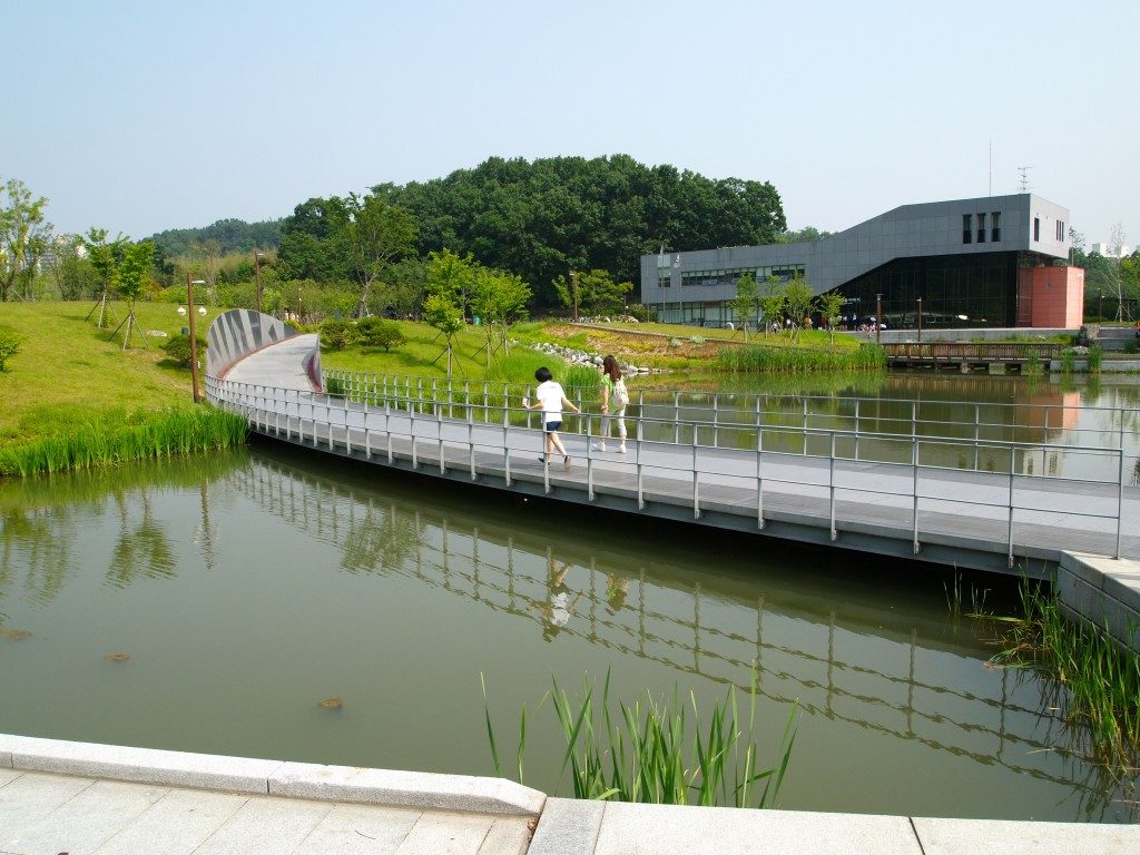 A pedestrian walkway crossing a river in Seoul's Dream Forest, with a brick and concrete building behind, two children on the bridge, surrounded by green grass and bushy vegetation.