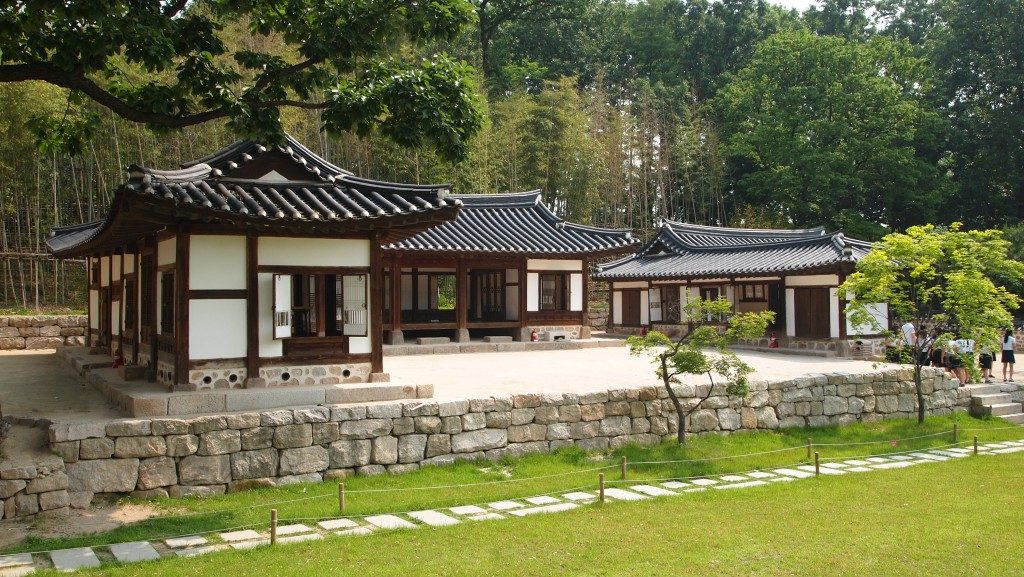 Exterior of the Dream Forest visitor center, a white and dark brown building with a courtyard in front, with stone walls, a stone path along the grass and thick forest behind.