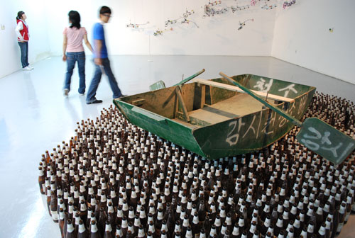 Exhibit at Gwangju biennale, a green wooden boat with oars, and white Korean lettering, sits atop a large number of bottles, arranged to look like the sea. In the background are people walking against a soft focus white wall.