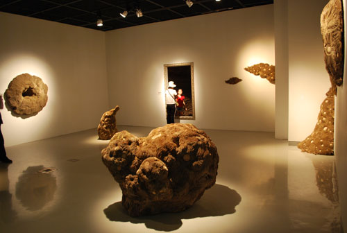 A darkened, spot-lit room contains an exhibit of dark brown rocks, on the wall and on the floor.