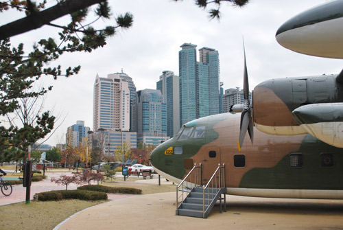 The front part of a fighter plane with steps up to the cockpit at Boramae Park, skyscrapers and roads in the background.