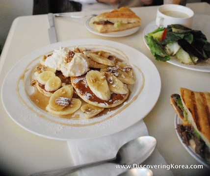 A white plate with pancakes, banana slices and maple syrup, topped with ricotta cheese, to the side is a brunch sandwich, and a plate of vegetables, on a white table.