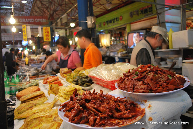 A stall vendor at Jungang market, with white plates of meat cooked in red spices, fried fish and condiments. In the background is the busy market, with people cooking and shopping.