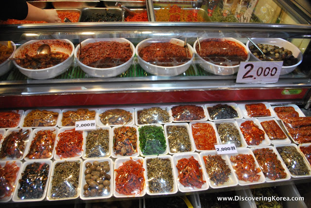 A glass cabinet containing different vegetables, herbs, spices and cooked meats, all in white trays wrapped in plastic.