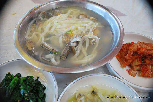 A metal bowl with a cloudy broth noodle dish, with clams and seafood. Three white plates contain condiments.