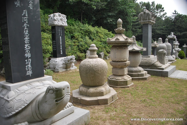 Stone plinths, on the left is a stone turtle, with a black headstone, with Korean inscription. In the background is a green hedge and grass.