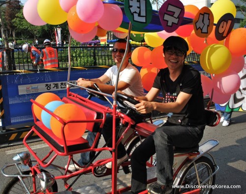 Two men on a bicycle rickshaw, with balloons drive through Seoul's streets at the Queer Culture parade.