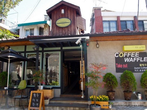 Exterior of a coffee shop in Seoul, a wooden building with seating outside, ornamental trees in pots, and a chalkboard sign with Korean lettering.