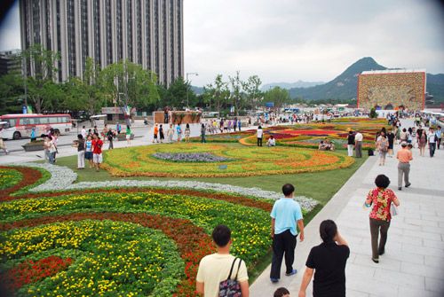 A large multicolored flower carpet as part of Gwanghwanun Square, circular plantings of different colored flowers with stone walkways down either side, with pedestrians, against a backdrop of a large building and a mountain in the distance.