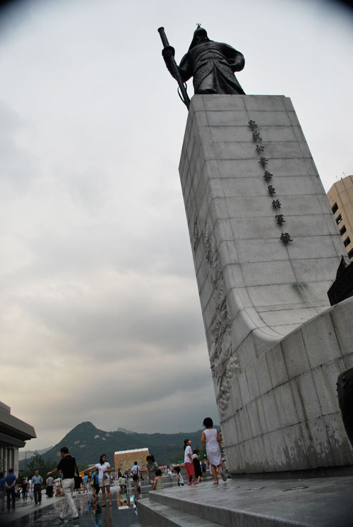 To the right of the frame is a tall concrete plinth with a statue on top of it, to the left are pedestrians on a street with no cars, the background is gathering clouds and a mountain in soft focus.