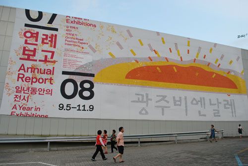 A large billboard, in white with red and black writing, and a red and orange picture, with pedestrians walking in the foreground.