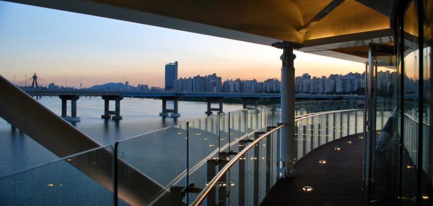 View across a river from a bridge with a glass and metal railing, towards the city of Seoul, in evening light.