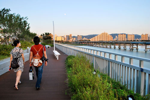 Two people walking over the Gwangnaru pedestrian bridge, with metal railings to the right of the frame, and some low bushes, to the left of the frame are trees and ahead is the city of Seoul in evening light.