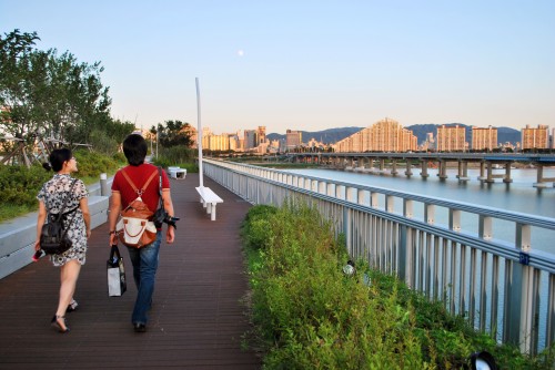 A man and a woman walking across Gwangnaru pedestrian bridge, with the river to the right of the frame, and cityscape behind.