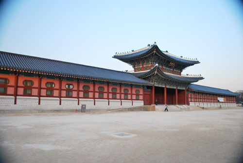 The light red exterior of Gyeongbokgung, showing an ornate two story entrance way, with dark roofing, and a stone colored area in front.