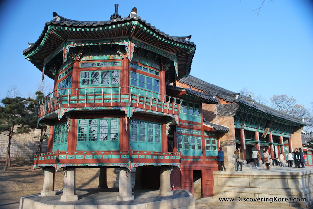 A view of Gyeongbokgung showing red and turquoise pillars and windows, with an ornate roof. To the right of the frame are a lot of visitors on a terrace outside.