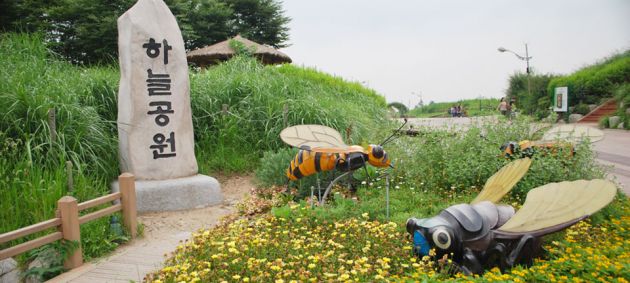 Two large sculptures of bugs in grass and wildflowers next to a stone sign with Korean lettering at the end of a footpath, with long grass in the background at Haneul park, Seoul.