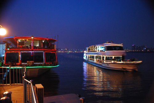Night view of two lit up boats on Hangang river, the left one has red and green lights, the right one has yellow lights. Soft focus view of the city lights in the distance.