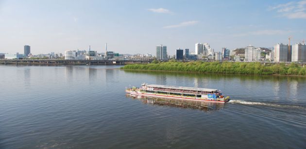 Hangang River with a boat in the foreground and cityscape in the background, with a blue sky and wispy clouds.