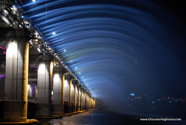 Hangang river at night, with the bridge to the left of the frame, and blue lights streaming into the river.