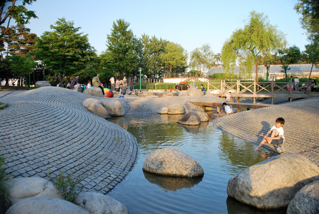 A water feature on Seonyudo island, showing paved banks going down to shallow water, with large rocks, children playing in the water, with trees and blue sky in the background.