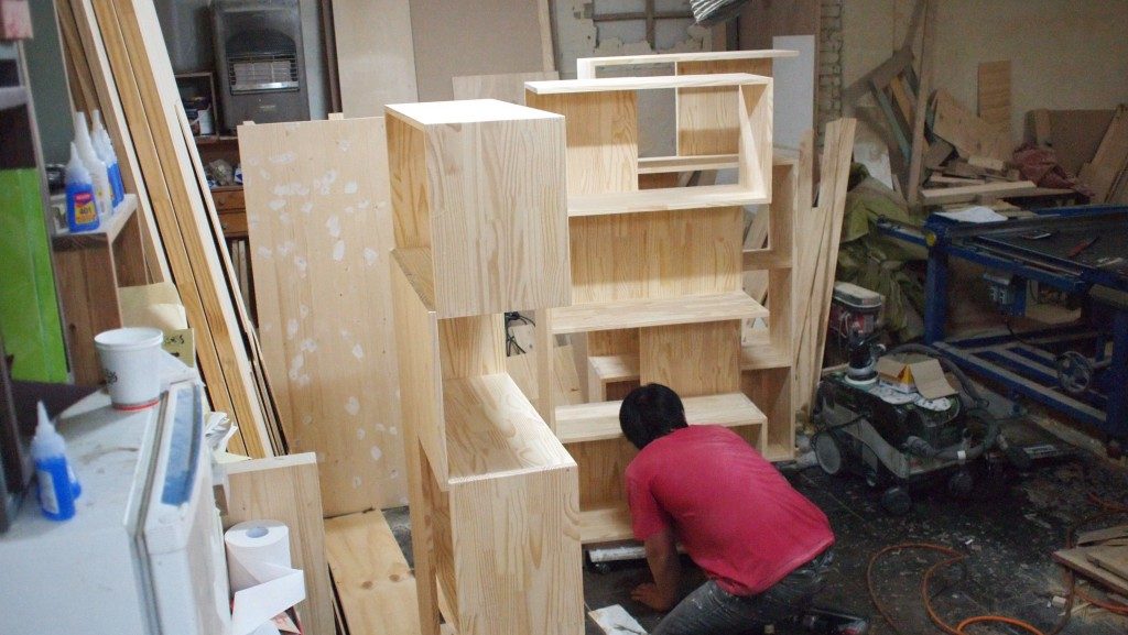 A man in a red shirt, kneeling down with a stripped pine cabinet he's working on in front of him. In the background are various tools and pieces of wood in the furniture workshop at Hongdae.