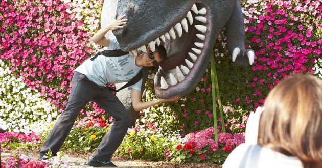 A young man leans into the mouth of a model dinosaur, putting his head between the sharp teeth, while a woman in the right of the frame takes a photograph. The backdrop is pink and white flowers in bright sunshine.