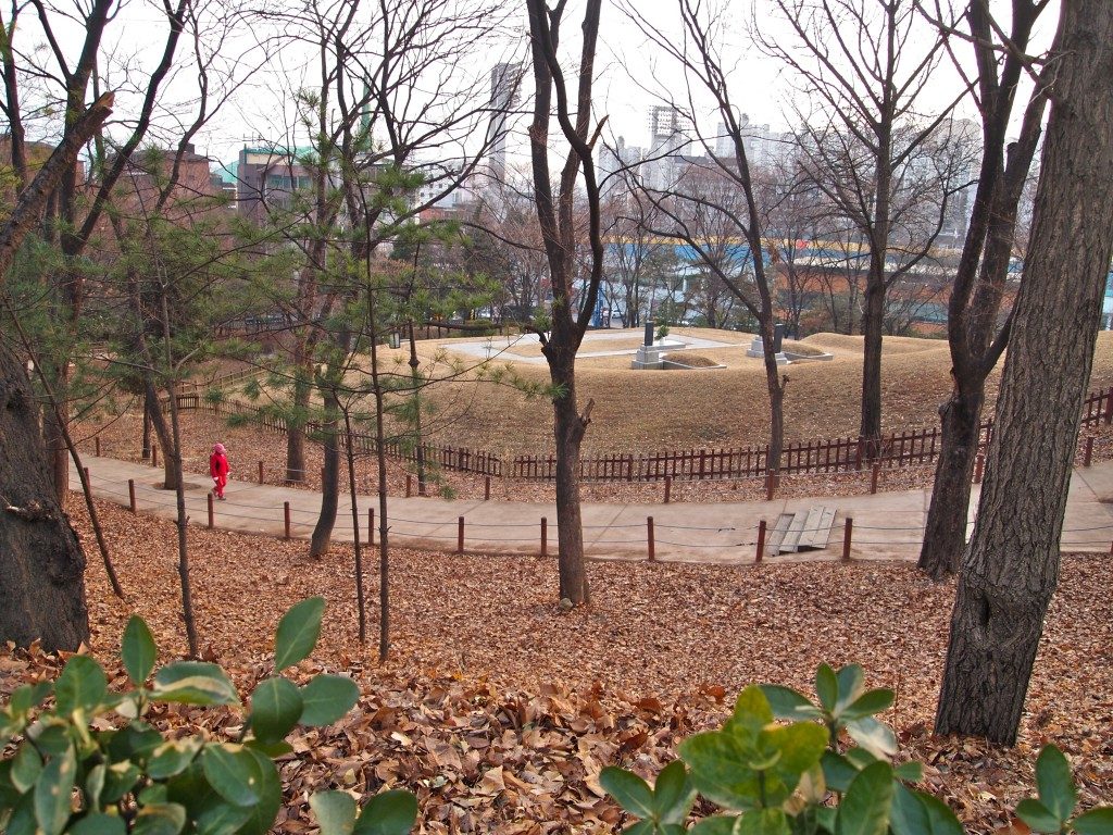 A view of Hyochang Park, with a pedestrian walkway, trees, and fallen leaves on the ground, with Seoul city buildings in the background.