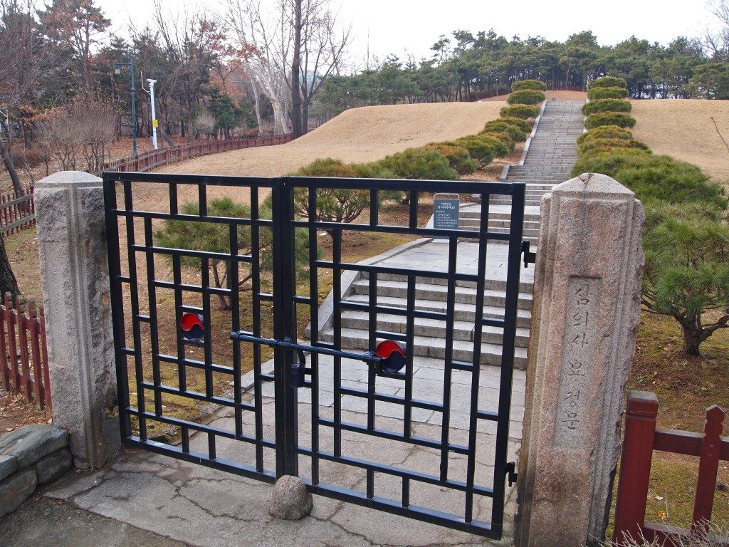 Black metal gates, set between carved stone posts at the entrance to Hyochang park, with stone steps leaving up in between low bushes with grass in the background.