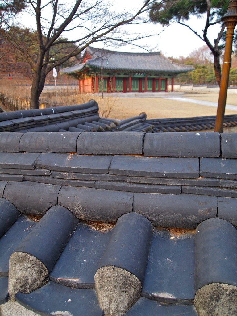 View over a tiled roof towards a shrine building, red pillars with green shutters, with vegetation and grass to either side.