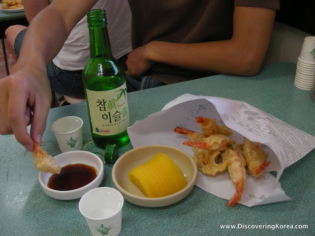A hand to the left of the frame dips a deep fried shrimp into soy sauce. A beer bottle, and paper packet of shrimp on a green table.