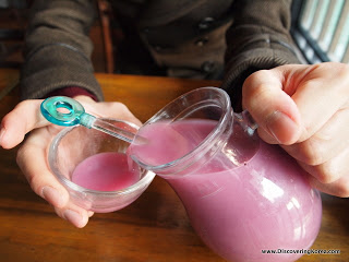 A hand pouring fruit infused rice beer from a jug into a small glass cup on a wooden surface.