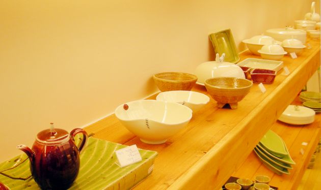 Wooden shelves containing ceramic items, some bowls, a tea pot, serving dishes against a yellow background.