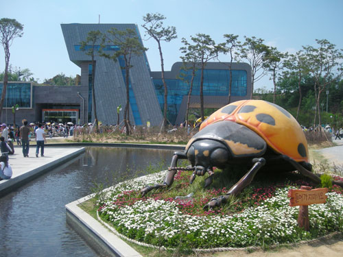 A large statue of a yellow and black beetle, surrounded by flowers, next to a water feature with a large stone and glass building in the background and pedestrians walking.