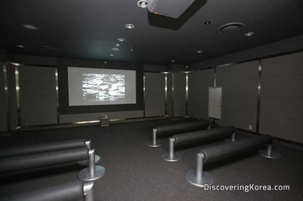 Interior of a screening room, dimly lit with a television on the far wall, and metal bench seating.