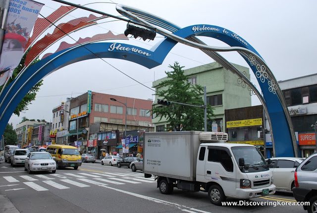 A street in Itaewon, Seoul, showing a blue and red archway over the street with cars and trucks and shopfronts in the background.