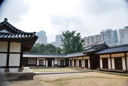 A traditional Korean building built around a courtyard, wooden frame and white walls, with a traditional curved roof. In the background are the modern buildings of Seoul.