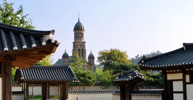 View towards the cathedral at Jeonju, in the background the cathedral has three dome shaped spires and arched windows. In the foreground are the traditional dark tile curved roofs of the hanok houses.