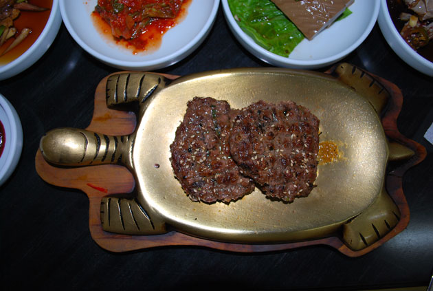 In the center of the frame is a brass plate in the shape of a turtle, with two beef patties, behind are four white dishes containing condiments.