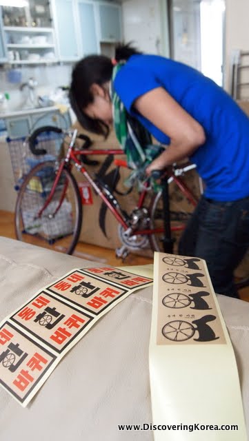 A woman using a hand pump to inflate a bicycle tire in the background. In the foreground are rows of printed stickers in black and red.