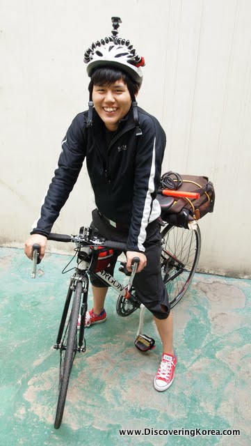 A woman pictured smiling at the camera on a bicycle, wearing a black outfit with a white helmet, on a pale green concrete surface.