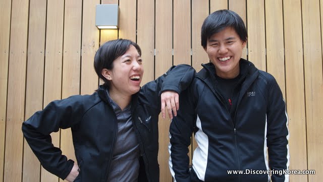 Two women pictured, one with her elbow on the shoulder of the other, both wearing black, against a wooden slatted background.